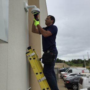 Electronic Security Technician on a ladder installing a video surveillance camera at an office building with vehicles in parking lot behind