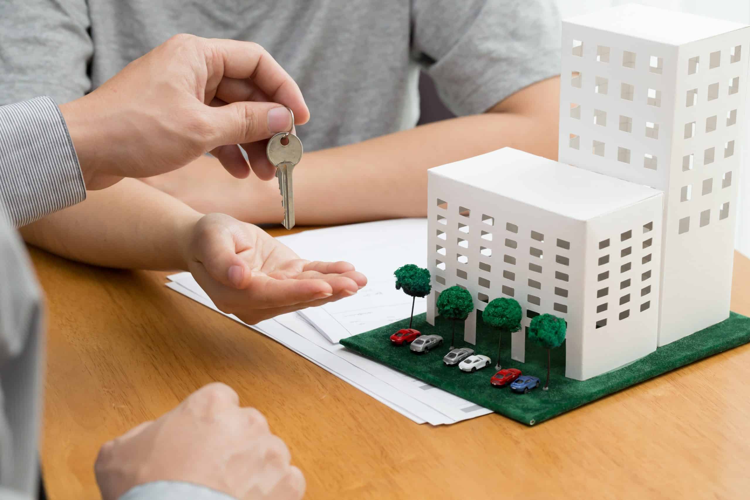 Person taking keys from another person beside a model condominium structure with formal documentation on a tabletop