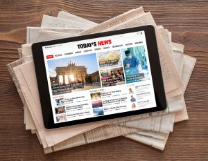 Tablet on stack of newspapers, tablet screen reads "Today's News" and features a number of news articles with text and pictures. Articles are not official news stories, they are intended as graphic display.
